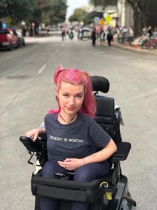 A white woman in a motorized wheelchair in the middle of an empty street. She has bright pink hair and is wearing a grey shirt with the words "This Body is Worthy" written across the front.